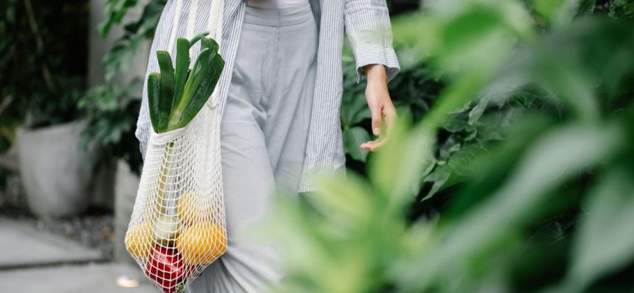 Crop anonymous female wearing casual outfit carrying net grocery bag with ripe vegetables including leek and capsicums in garden