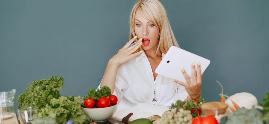 A surprised woman looking at a tablet surrounded by fresh vegetables and fruits, promoting healthy eating.