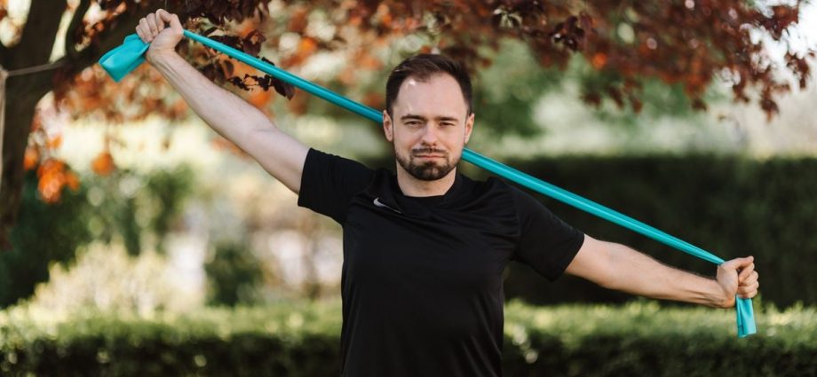 Adult man in black shirt exercises with a resistance band outside.
